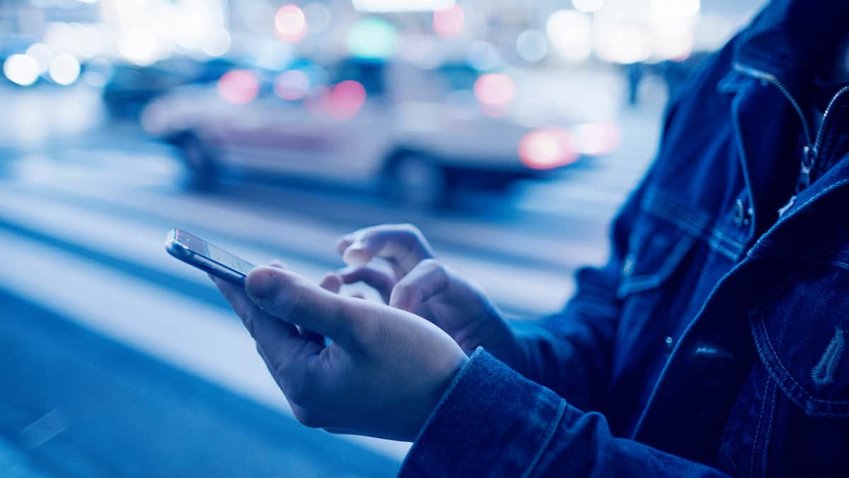 A person taps their smartphone outside on the sidewalk where the street lights and cars are motion blurred