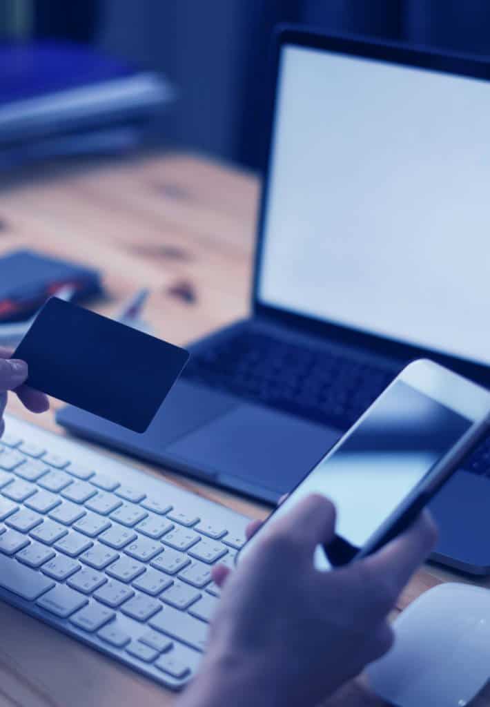 A person holding both their smartphone and credit card in their hands with a laptop on a wooden table in the background