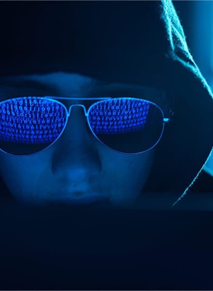 A man in sunglasses and a hoodie stares at lines of binary on his laptop screen.