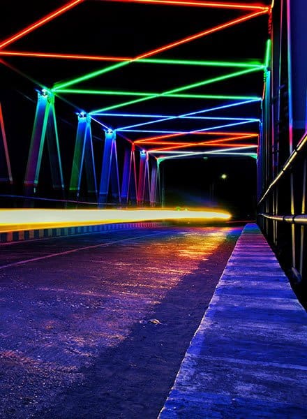 Sidewalk and road on a bridge with a light blur and colorful lights criss crossing as the top of the bridge