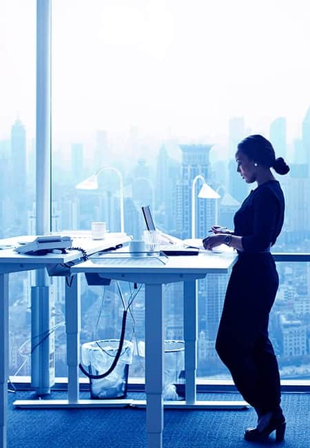 A working woman stands at a standing desk in front of a wall made of windows