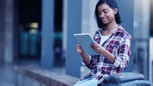 Girl on her tablet sits on a bench with her backpack by her side