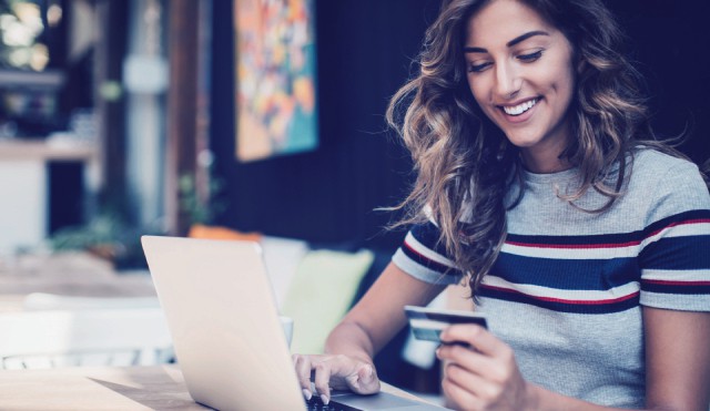 A woman smiles as she types her card information on her laptop, presumedly to complete an online purchase.