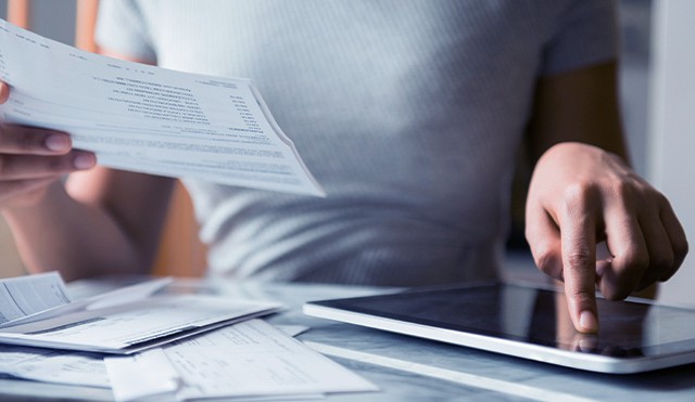 A woman holds a bill while scrolling on her tablet.