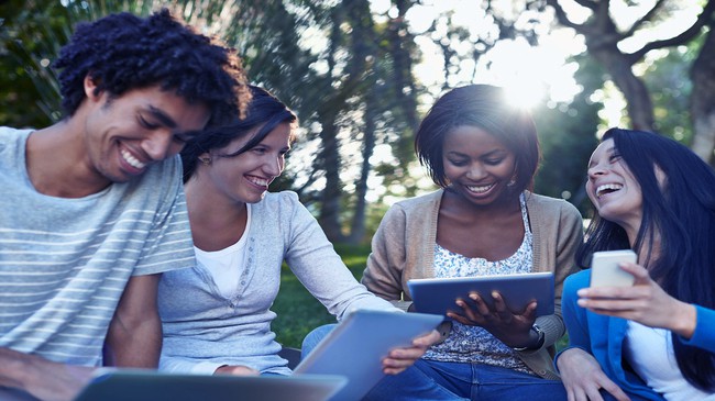 A group of friends sit in a park each has a different smart device as they laugh together.