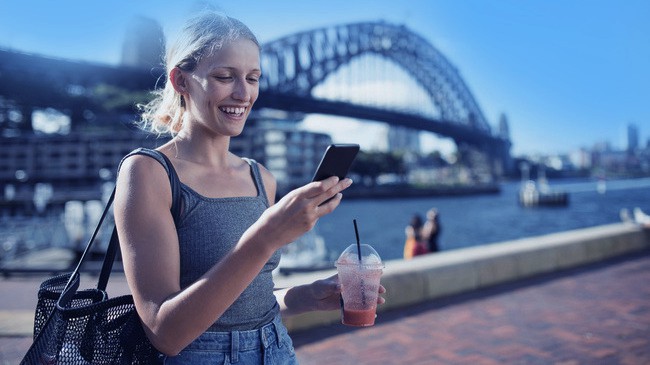 A woman holding a smoothie on the waterfront smiles at her smartphone with a large bridge in the background