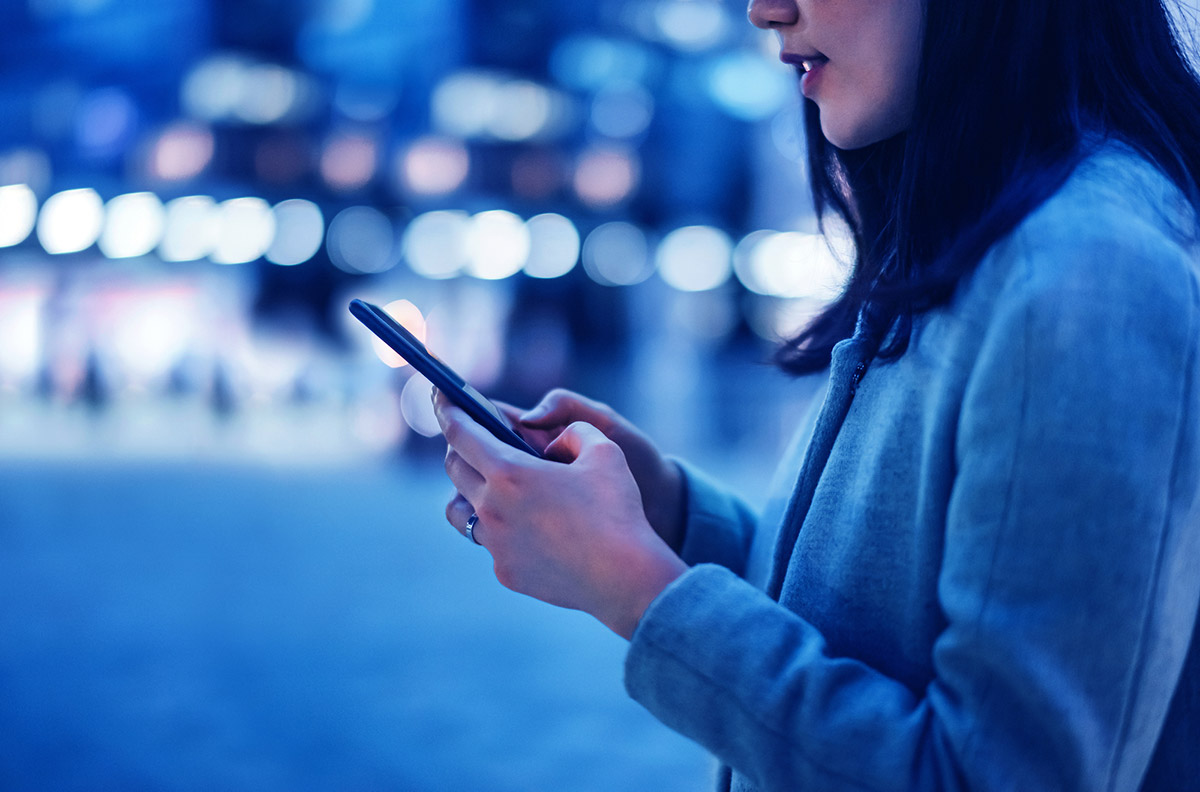 A woman is standing on the beach at twilight, she is on her phone while there is a bokeh of city lights behind her.