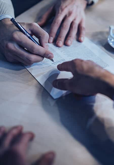 Two people sit at a table. There is a contract between them. One of them pointing at it and the other ready to sign.