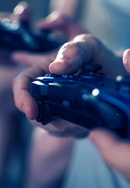 A man holds a playstation controller, there is another person in the background holding a controller as well, they are playing a video game together. 