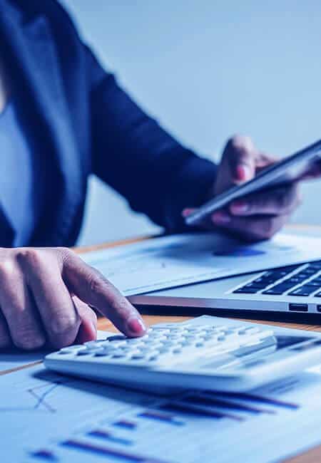 A person is using a calculator on their desk filled with papers, there is a smartphone in their other hand.