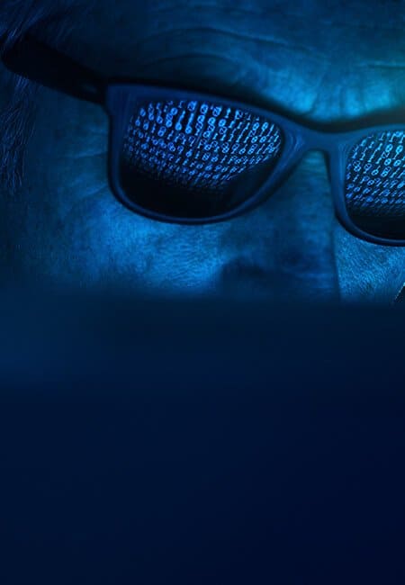 An old man sits in front of a computer screen in the dark, he is wearing sunglasses and in the reflection of these glasses there are lines upon lines of code.