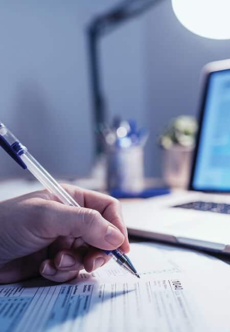 A person is filling out their taxes at their desk with a pen and paper, their laptop is in the background.