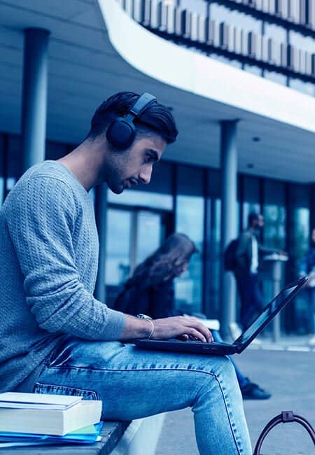 A man sits on a bench in public with headphones on, there is a stack of books next to him while he types on his laptop. 