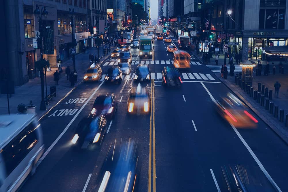 Photo of a multi lane street in a busy city. There are people walking on the sidewalks and cars driving by with motion blur.