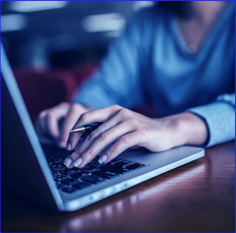 A woman with a French manicure holding a pen is typing on her MacBook Pro while sitting at her dark wooden desk. She is wearing a gray sweater.