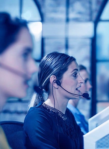 A woman at a call center is actively talking to a customer over the phone with two of her coworkers in the picture also talking to customers. One is in the foreground and one is in the background, the only one in focus is the woman in the middle.