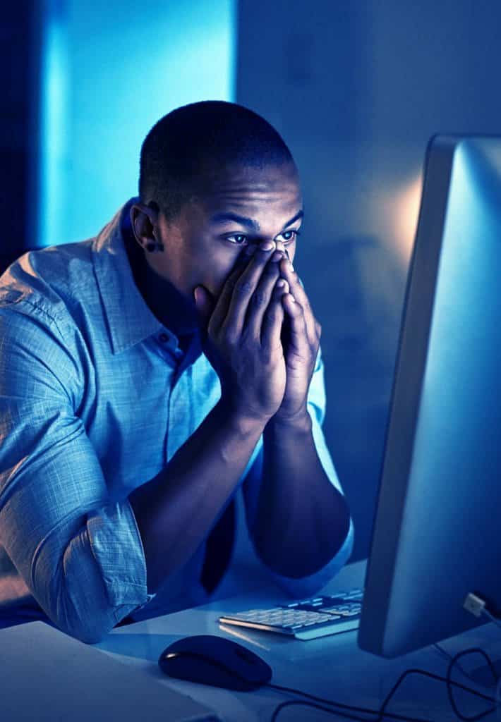 A man sits at his computer screen in a dimly lit room. His hands are covering his face in either contemplation or frustration.
