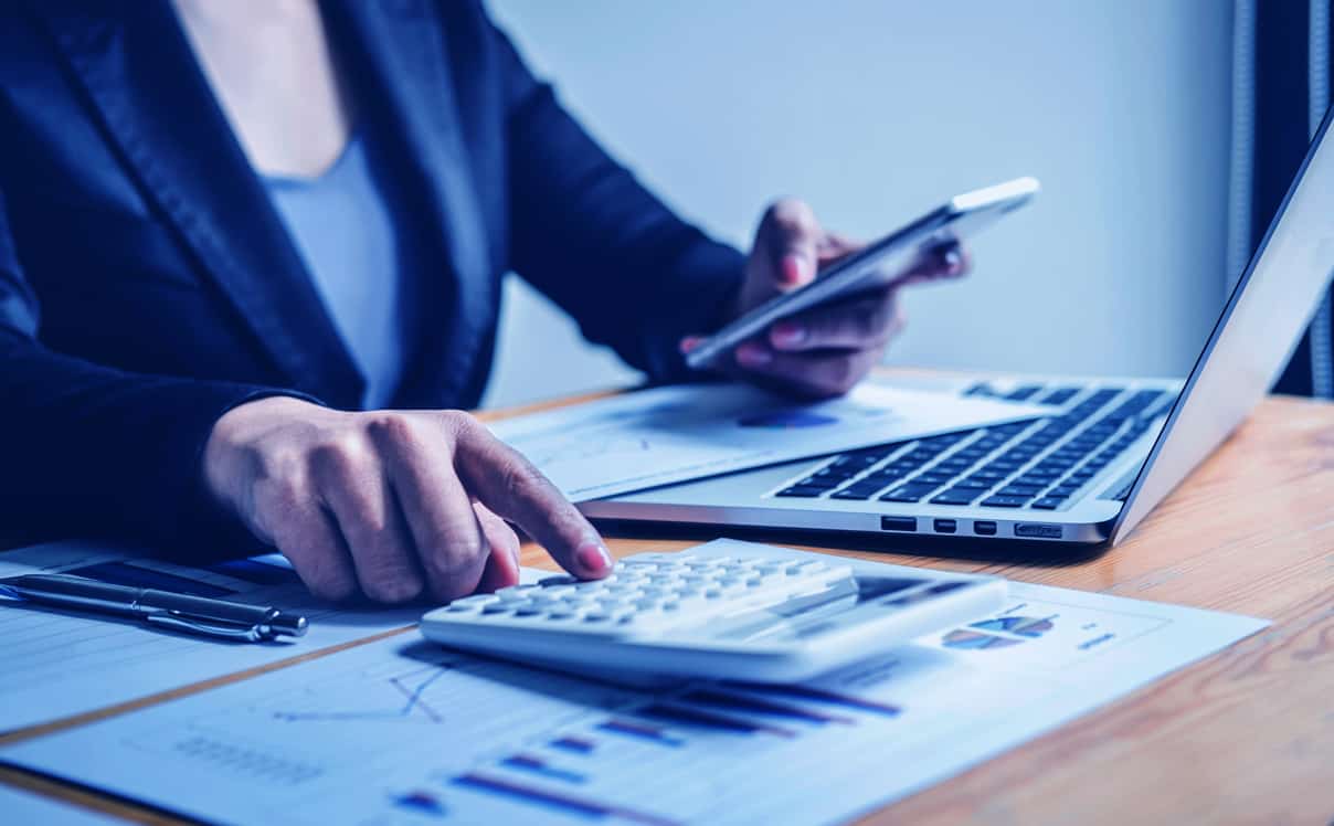 Business woman sitting at her desk holding her smartphone while using a calculator with reports and her laptop in front of her