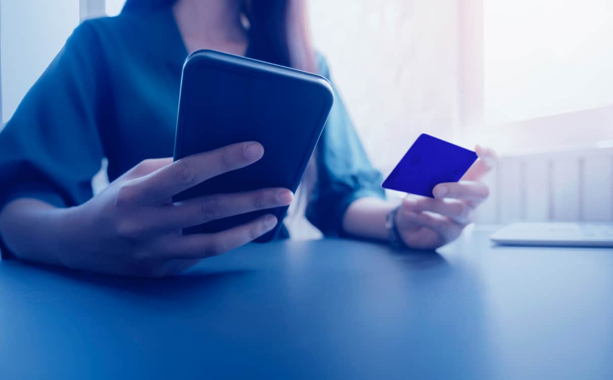 A woman holds her phone in her right hand and credit card in her left hand, she is paying for something on her phone.