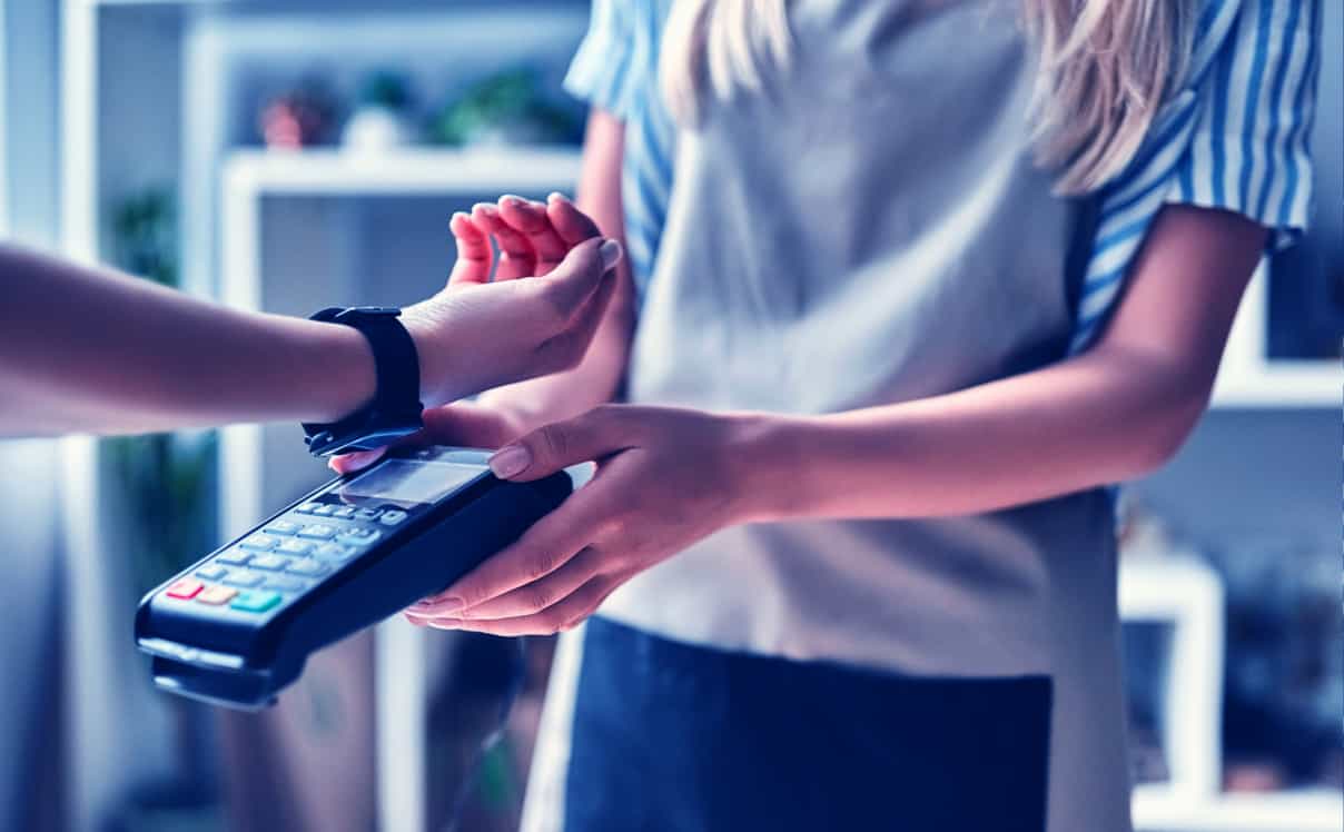 A woman uses her smartwatch to pay for her transaction with a tap to pay point of sale system.