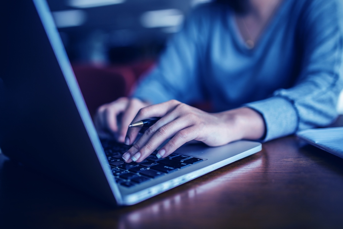 A woman with a French manicure holding a pen is typing on her MacBook Pro while sitting at her dark wooden desk. She is wearing a gray sweater.
