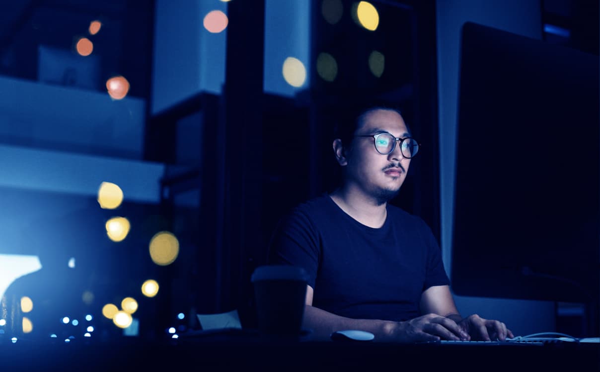 A man sits at his desk in the dark with his computer in front of him, there is a large glass window with reflecting lights behind him.