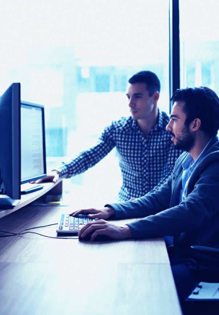 Two men are at a desk starting at two monitors, they are working on a project together.