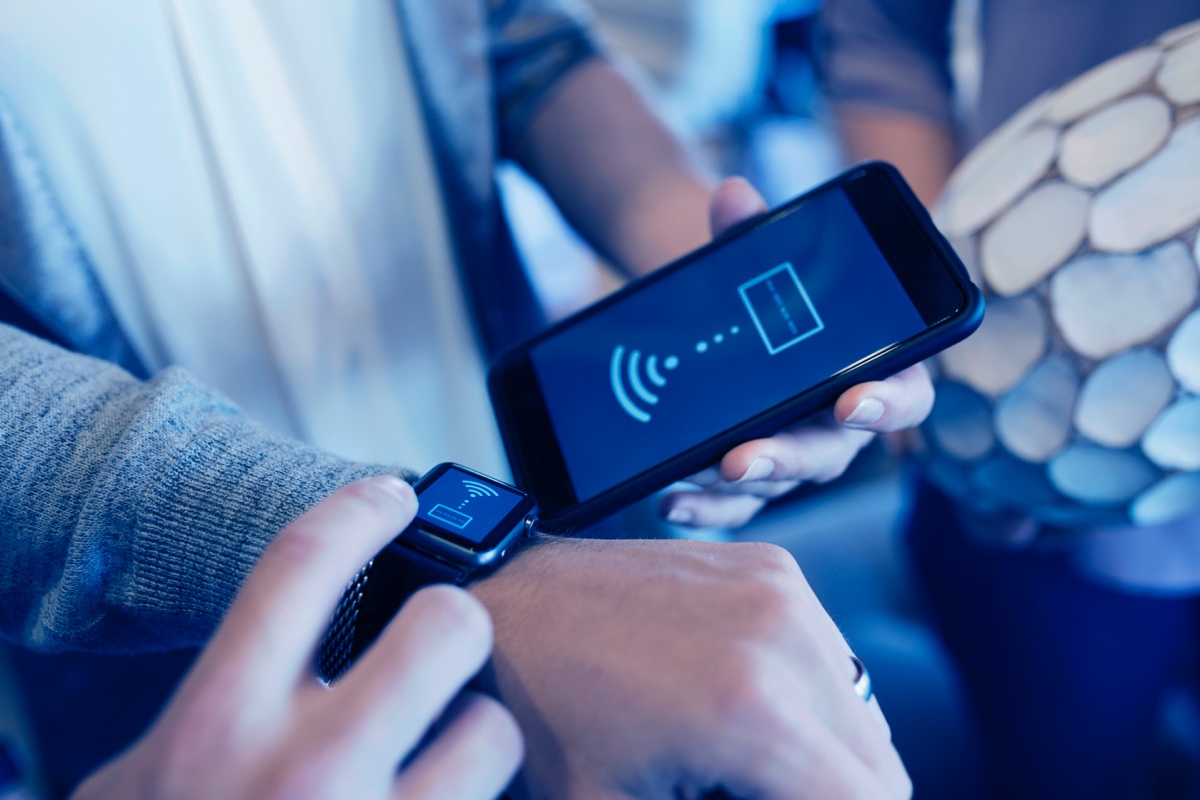 A woman uses her phone to transfer funds to her friend by tapping her phone to their friends smart watch.