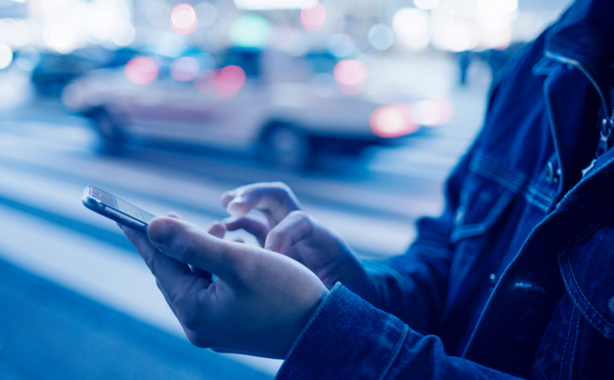A person is standing at a crosswalk using their smartphone, their are cars in the background and the image has a blue tint.