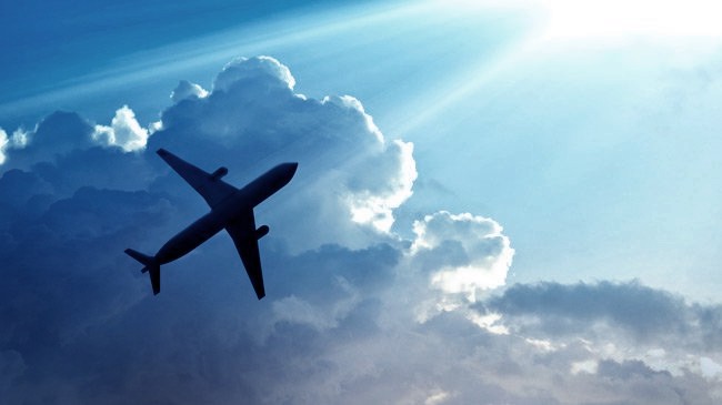 An upwards facing shot of a plane flying across the sky with a large fluffy cloud above it.