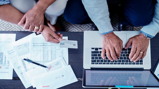 A person holds out their card while another man types the information out on their macbook pro, there are accounting sheets to the left that the two are going over.