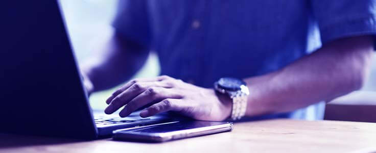 Man with a watch on types on his laptop with his phone next to him