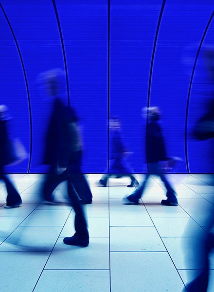 People walking past the camera in a tunnel, there are blue curved ceilings and a white tile floor. 