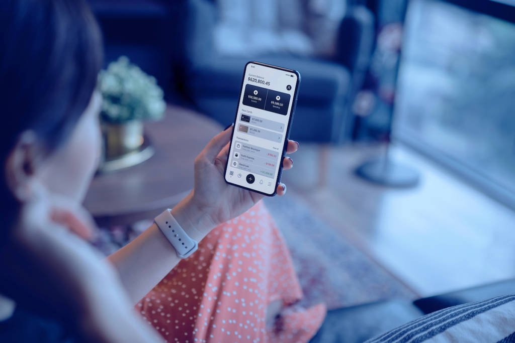 Young woman sitting on the sofa in the living room, managing online banking with mobile app on smartphone. Transferring money, paying bills, checking balance.