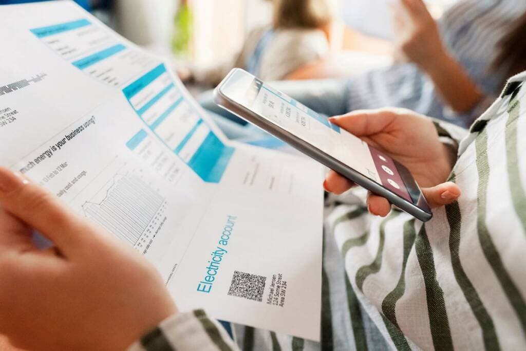 Person paying electricity bill with smartphone and printed bill in hand.