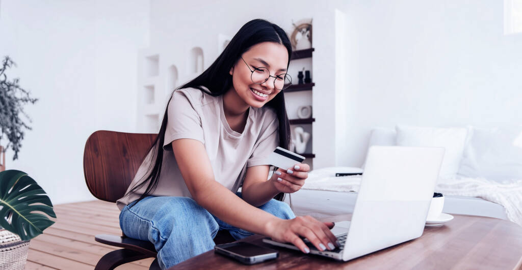 Woman using a laptop and smartphone at home, smiling and engaged in online activity.